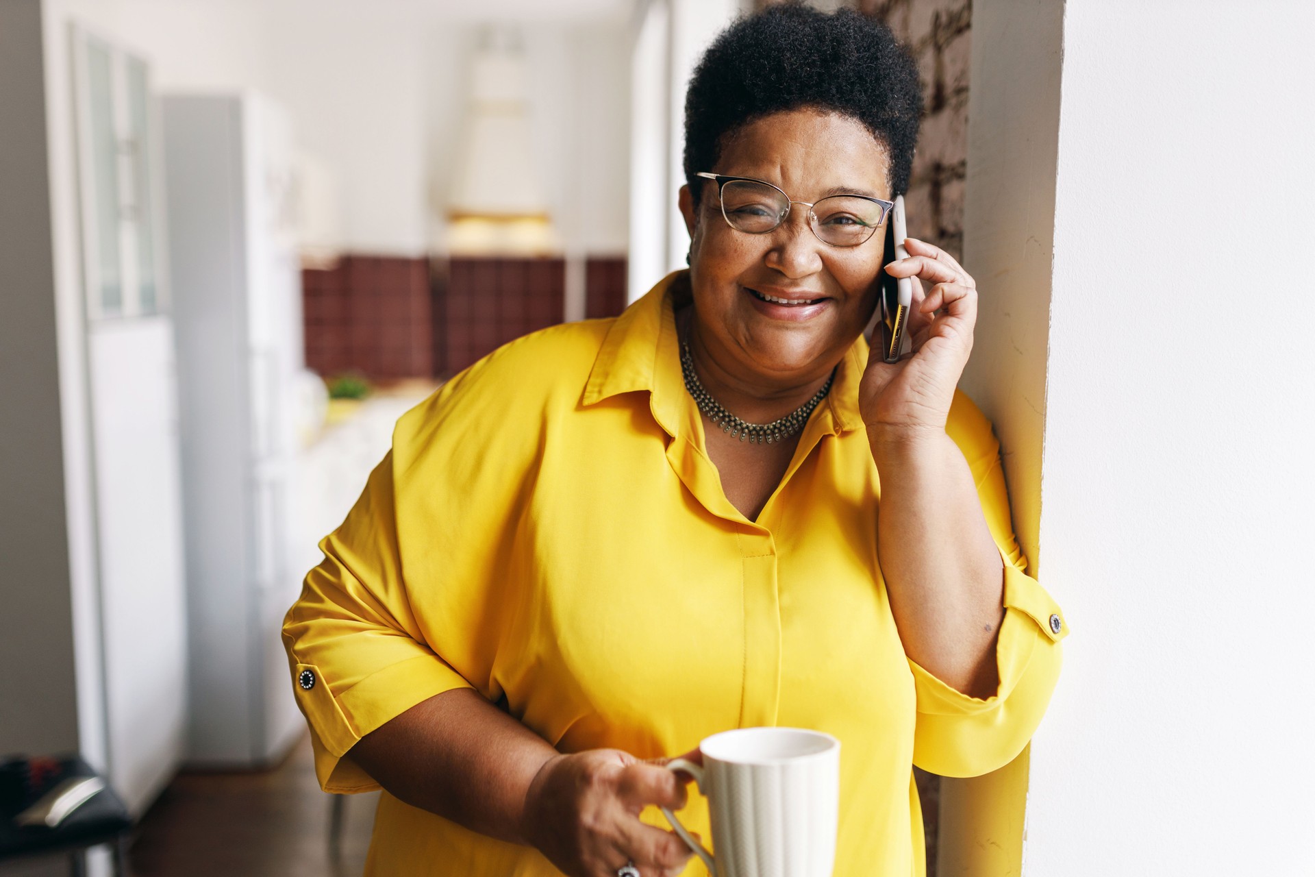 appy cheerful black senior plus size female with short haircut and eyeglasses having coffee break holding white cup while talking on phone with her friend, joking discussing funny situation